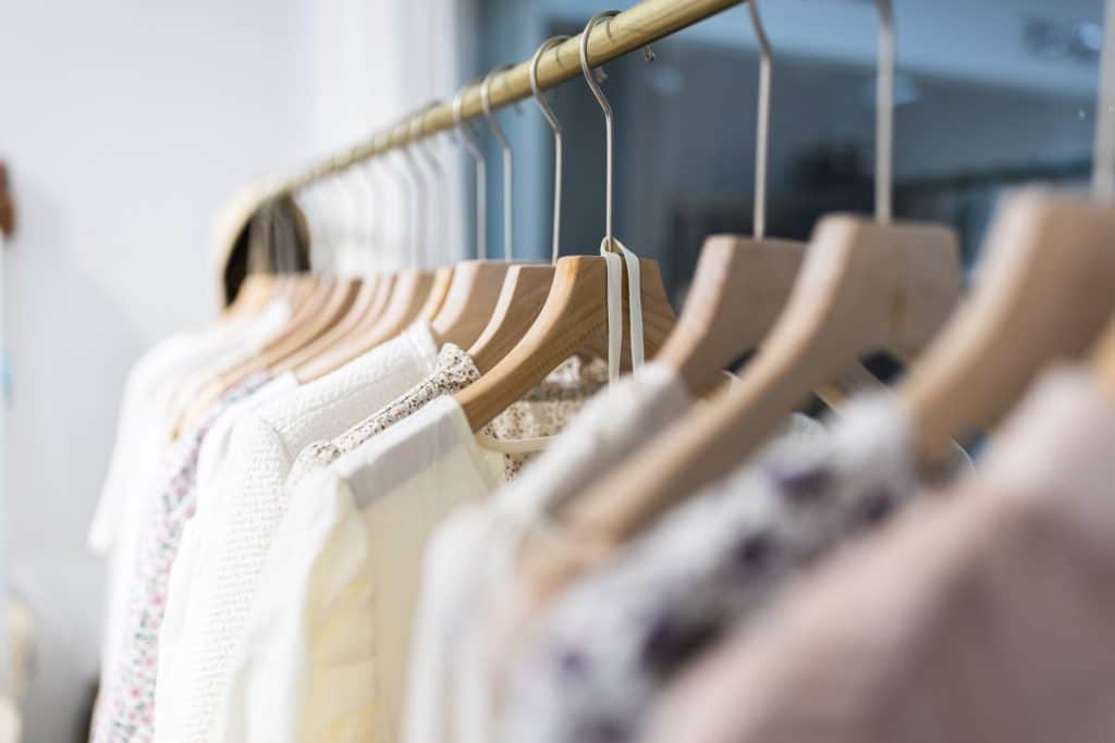 Perchero con prendas de ropa en tonos claros colgadas en perchas de madera dentro de una tienda.