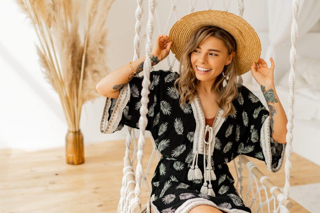 Mujer joven sonriente con sombrero de paja y vestido negro estampado de hojas blancas sentada en un columpio de cuerda.