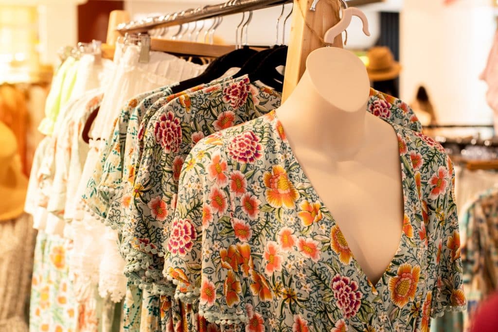 Vestido floral en tonos verdes, naranjas y rosas en maniquí, junto a prendas similares en perchero dentro de una tienda.