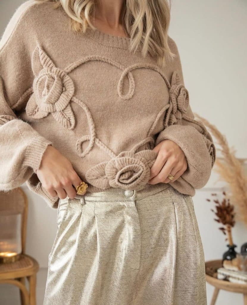 Mujer con jersey de punto beige con apliques florales y pantalones dorados de tejido brillante.