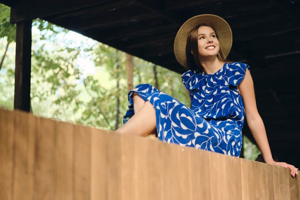 Mujer joven con vestido azul estampado y sombrero de ala ancha sentada en un entorno natural.